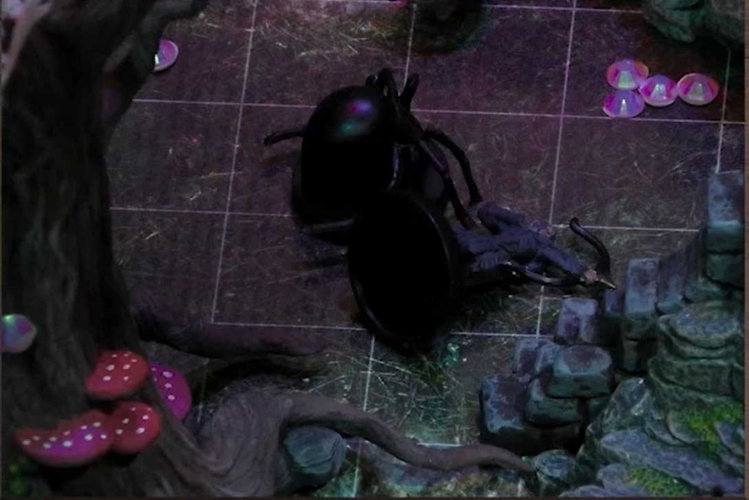 A battlemap of a forested area. A giant spider stands over a fallen Cyrus. Above them rises a leafless tree with red mushrooms growing from it. A group of crystals in the vague shape of a penis is on the ground nearby.