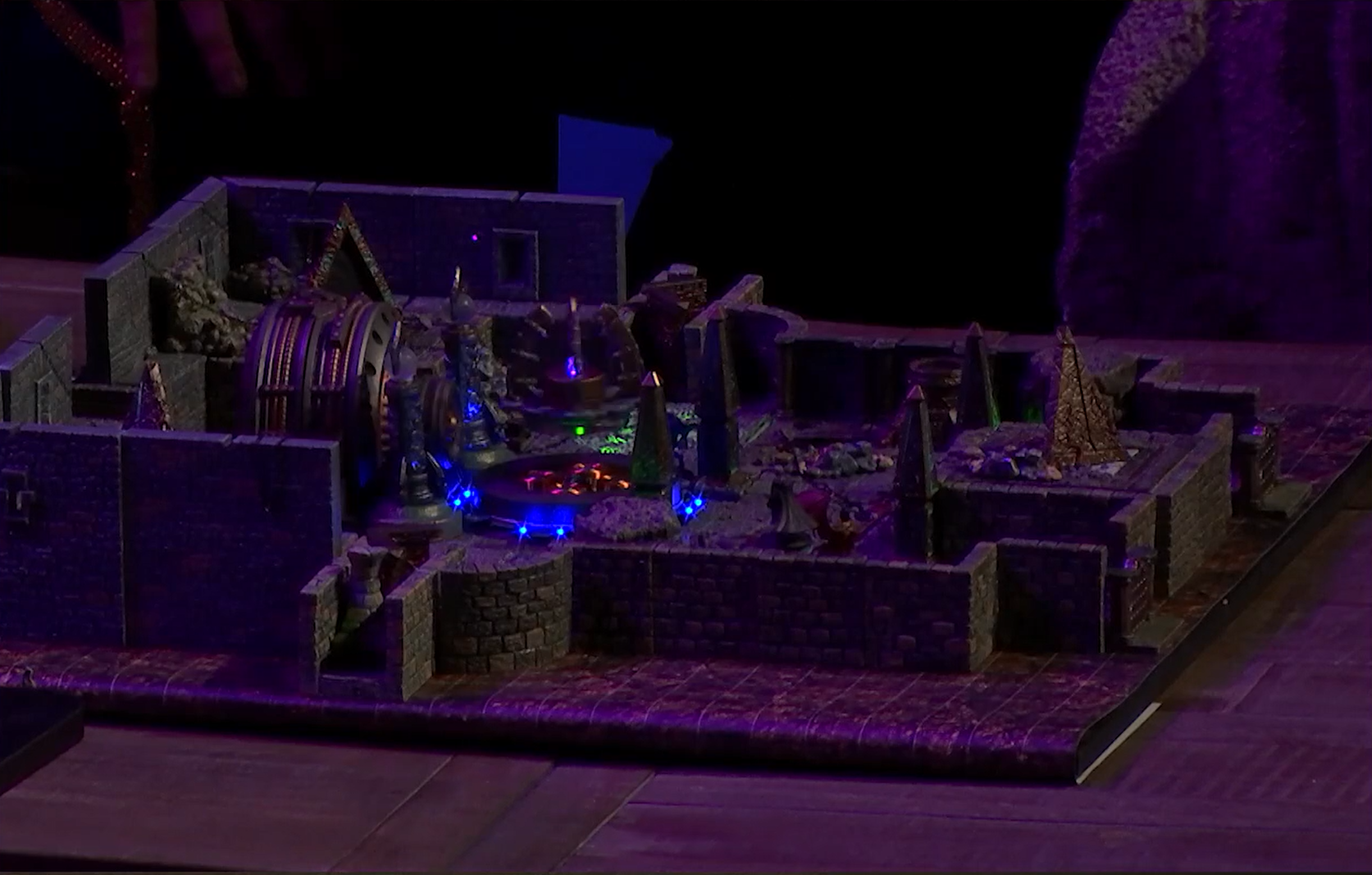 A battlemap of an indoor space with stone brick walls. To the left is a black and gold cylindrical machine with segmented tubes running along its circumference. In front of it is a floating platform on which are four inward-curving spires and a purple crystal. The platform floats red glowing lights and is surrounded by blue lights on the ground. Further to the right are four pillars surrounding a more open area of floor.