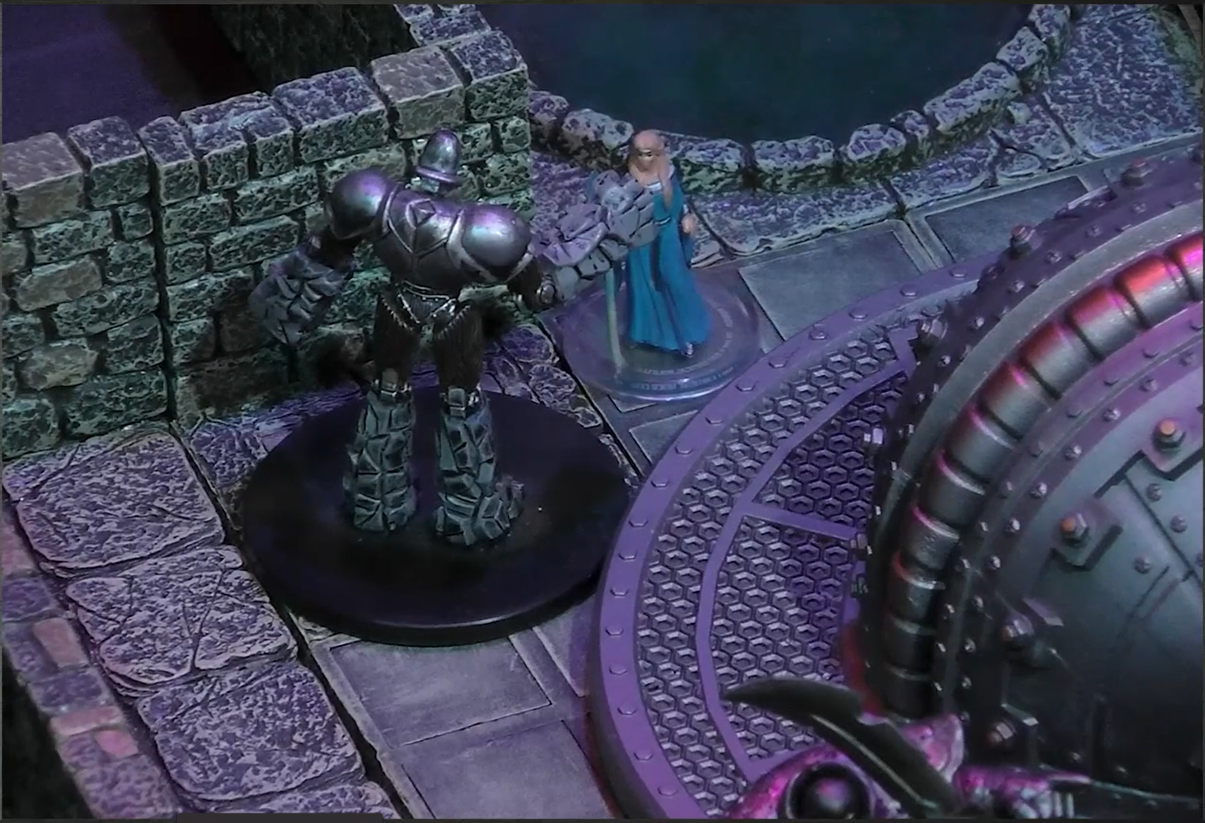 A battlemap of an indoor space with stone walls. The Emissary faces off against an archmage with a sky blue cloak and long blond hair. A dome-like metal structure is off to the side in the center of the room and a pool of inky shadow is in the back of the room.