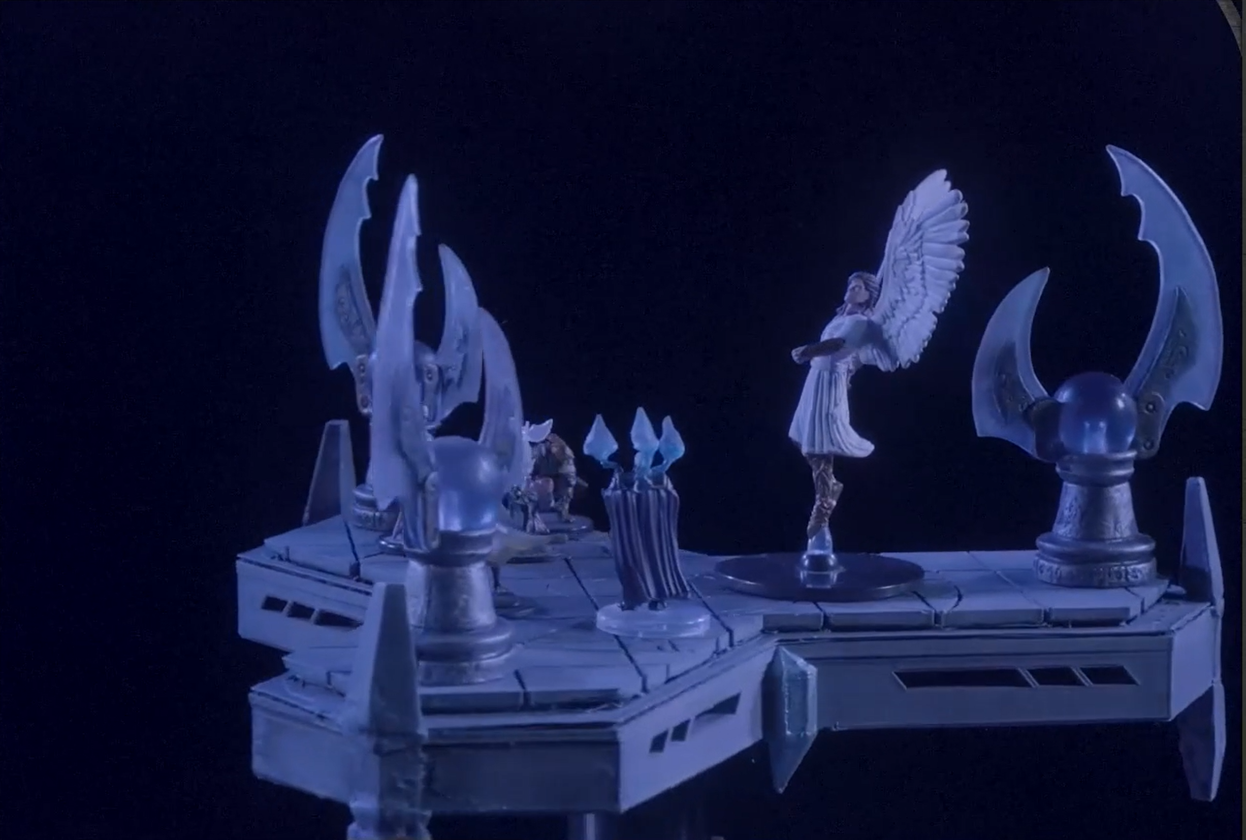 A close-up of a battlemap. An elevated three-tipped metal platform. The tall angel-winged Solar Acastriel stands to the side near two smaller figures. At the end of each tip of the platform is a ward, a globe on a pedestal with protruding translucent blades.