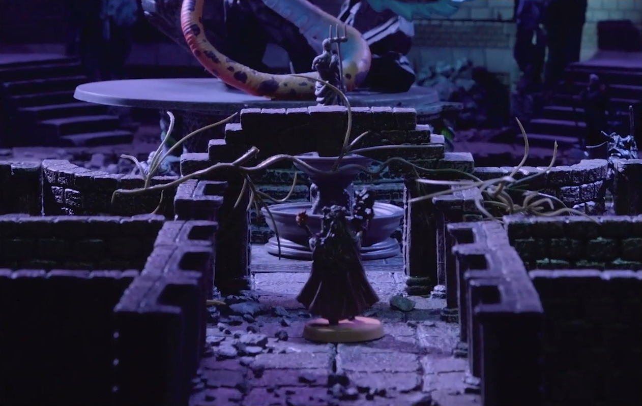 A battlemap looking down a hallway over a minifigure of Fearne. Past her are some vines stretching across the path. Beyond those are a fountain, a statue wielding a trident, and the red- and purple-spotted tail of the dragon Gloamglut.