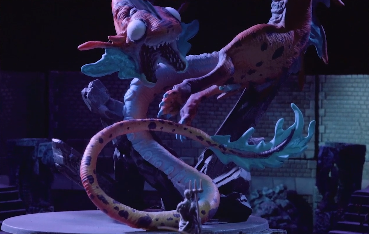 A battlemap focused on Gloamglut, a massive snake-like dragon. The large, blank white eyes of its fish-like head look forward, its split lower jaw open and filled with sharp teeth. Its rear legs trail not far below its head and above its sky blue frilled tail. A statue wielding a trident stands in the foreground.