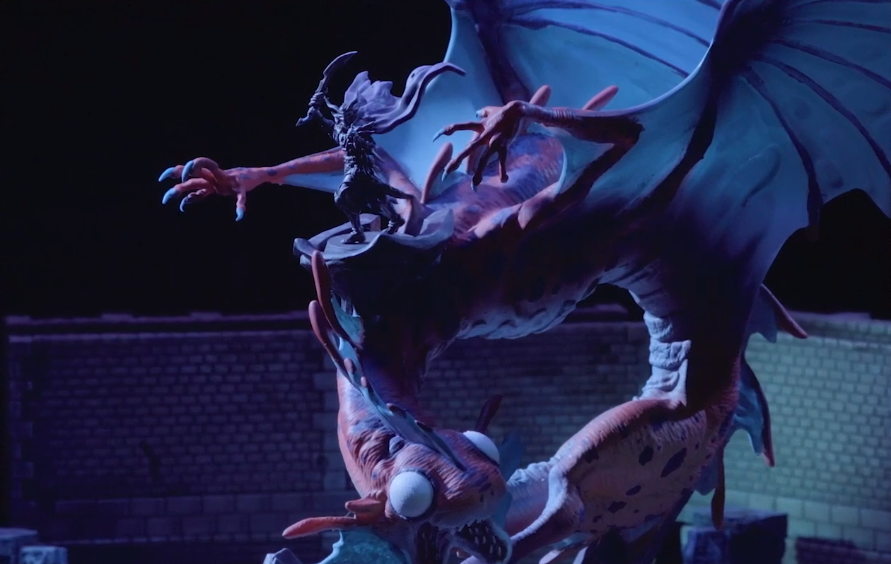 A miniature of Gloamglut, a massive snake-like dragon, stands in the middle of a stone chamber. Upon its back is Sorrowlord Zathuda in black armor holding a sword aloft. Gloamglut’s wings are outstretched. The large, blank white eyes of its fish-like head look forward, its split lower jaw open and filled with sharp teeth.