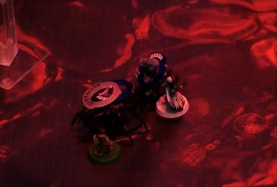 A battlemap with a glowing red blood floor focused on miniatures for Orym, Opal in spider form, Chetney in wolf form, and Braius. Opal stands between Orym, Chetney, and Braius, a humanoid torso emerging from the abdomen of a giant spider. Her miniature has rings on it labeled “HEXED” and “RESTRAINED”. Chetney’s has a ring labeled “HEXED”.