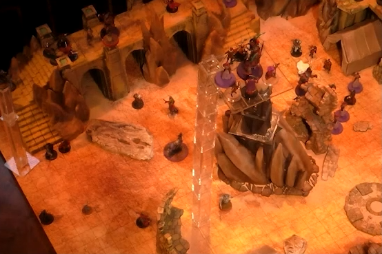 A battlemap of a large outdoor space filled with stone walls and parts of buildings. In the center is a tower with a pale white globe at the top. A number of combatants fill the area on the ground and atop the walls. A set of tokens is floating high above the battlefield.