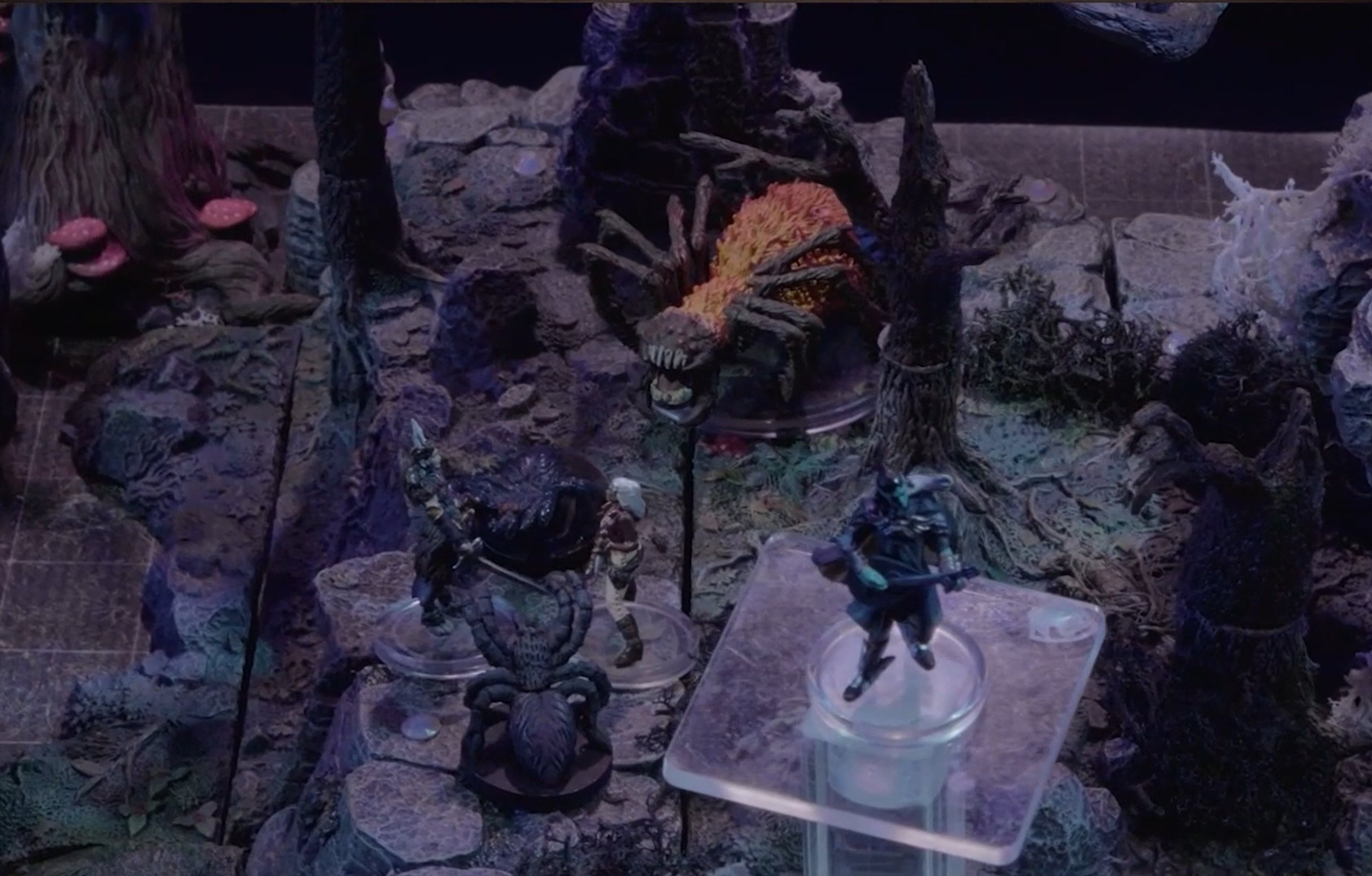 A battlemap of a rocky, forested area. Opal stands near Morrighan and a giant spider, while a massive spidker-like creature with a furry orange abdomen stands a little bit back. Dorian levitates in the foreground.