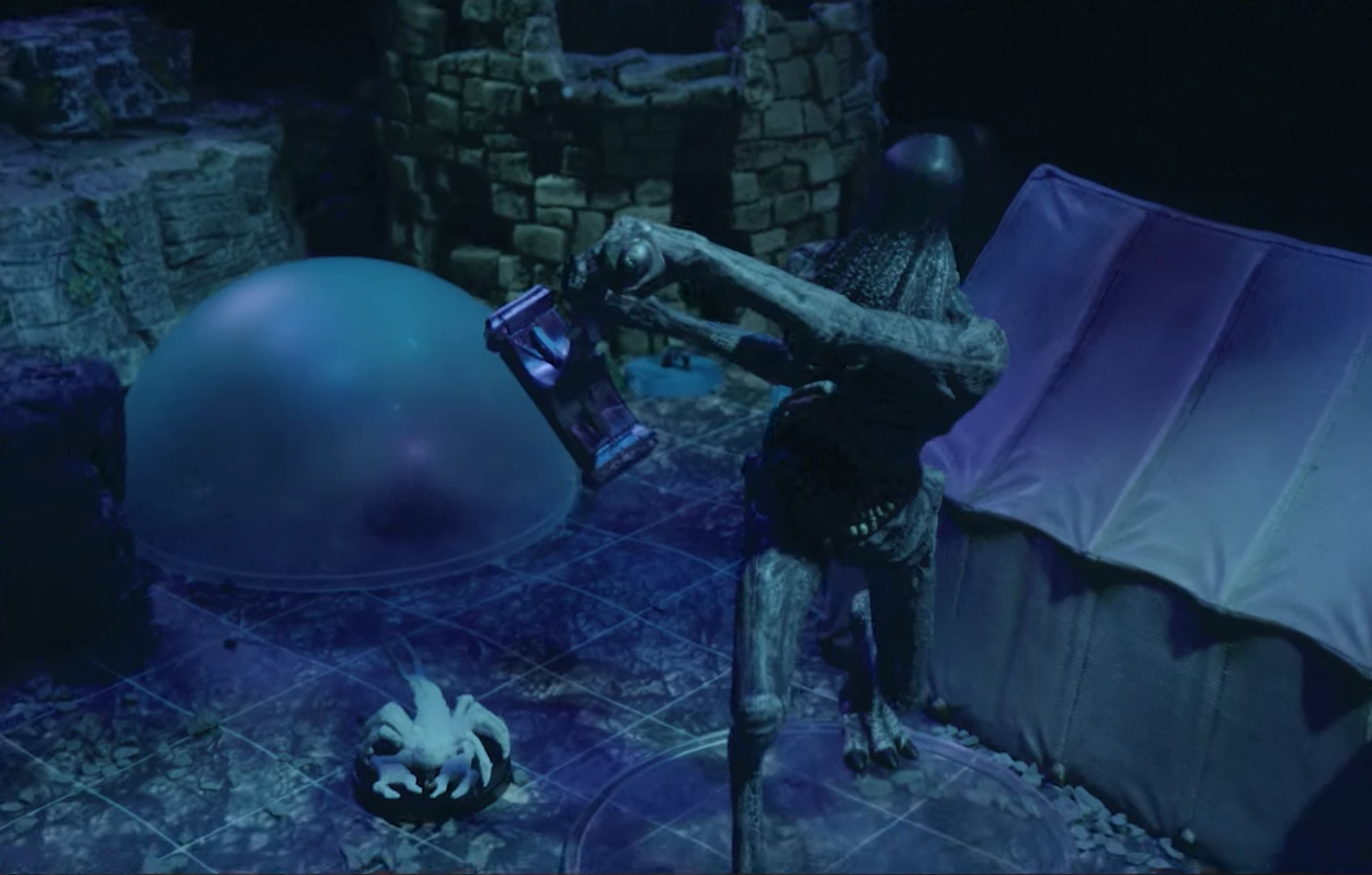 A battlemap focused on a Tomb Tapper. A tall, thin, gray creature with folds of skin hanging from its face swinging a hammer towards the camera. On the creature’s chest a toothy mouth is visible. The creature has feet with three toes ending in claws. In the background is a pale white scorpion, a square tent, and a blue translucent dome.