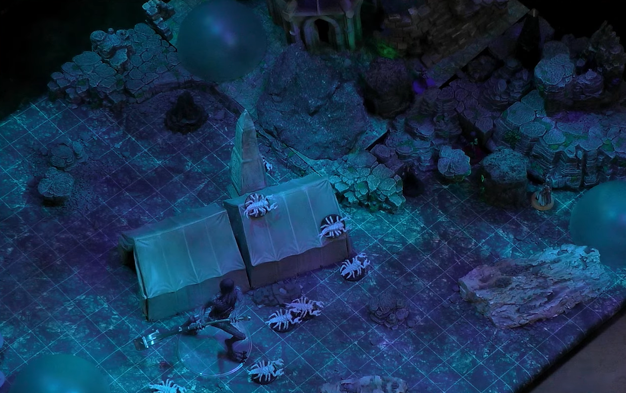 A battlemap of a dark underground cavern filled with the ruins of a civilization. In the center next to a pale white obelisk and two rectangular tents is a Tomb Tapper, a tall, thin, gray creature with folds of skin hanging from its face wielding a hammer. On the ground and tents near the Tomb Tapper are eight pale white scorpions.