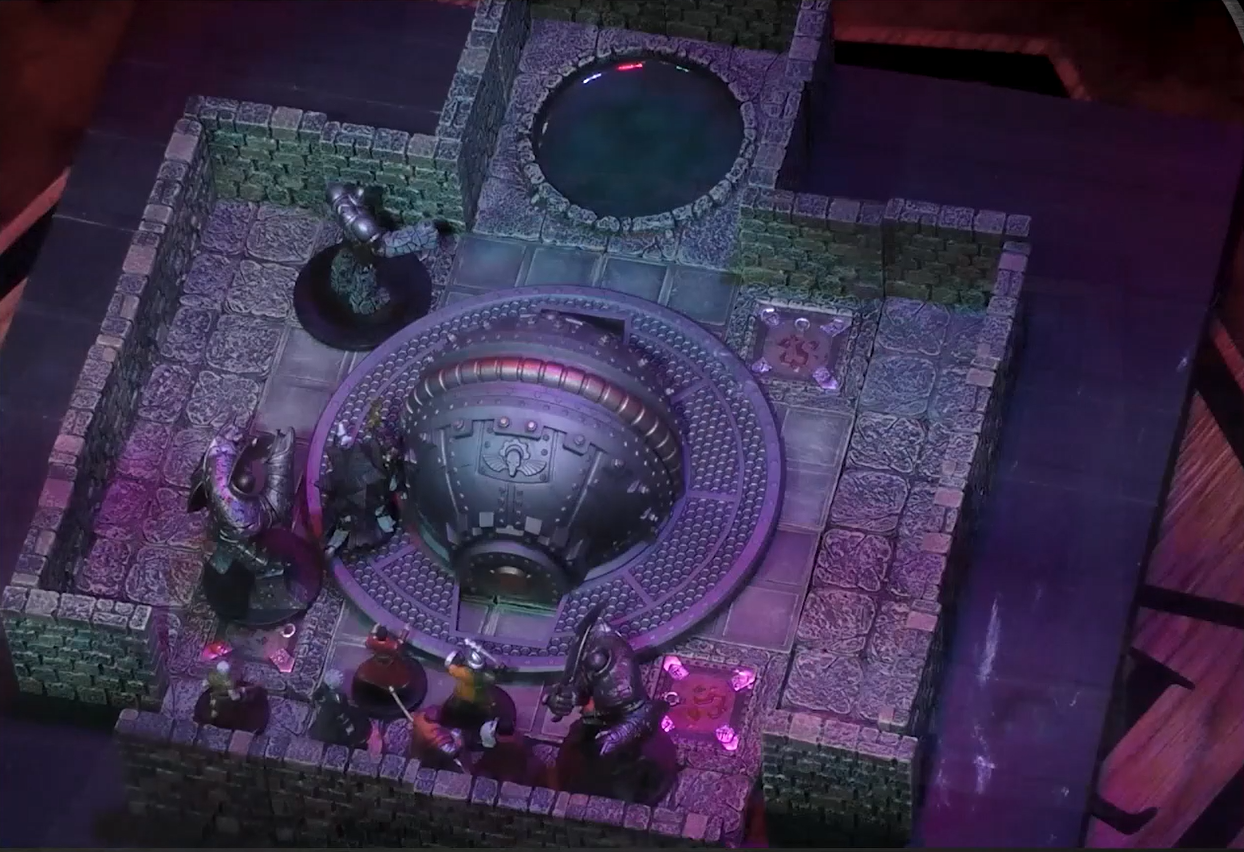 An overhead view of a battlemap of an indoor space with stone walls. A dome-like metal structure is in the center of the room and a pool of inky shadow is in the back. The Emissary is towards the back, with the rest of the party near the entrance facing off against two large metal humanoid constructs wielding curved steel blades. Zaharzht stands by the dome.