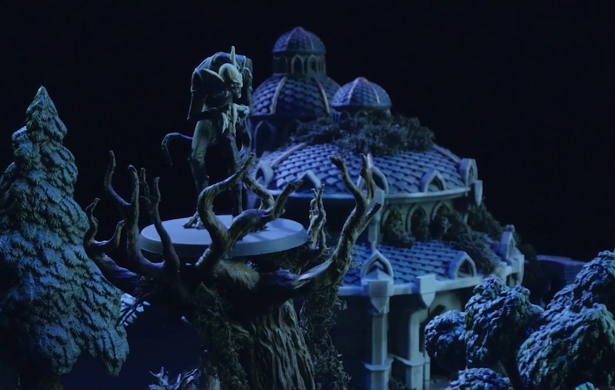 A battlemap focused on the Nightmare King standing at the top of a leafless tree. His pale form is tall and hunched over, his hands meeting in front of him with long fingers drooping down. Two long pointed ears sprout from his bald head and his bottom jaw is lowered into an open-mouthed sneer. Behind him is a large building with a three-tiered pale purple shingled roof and cupola.