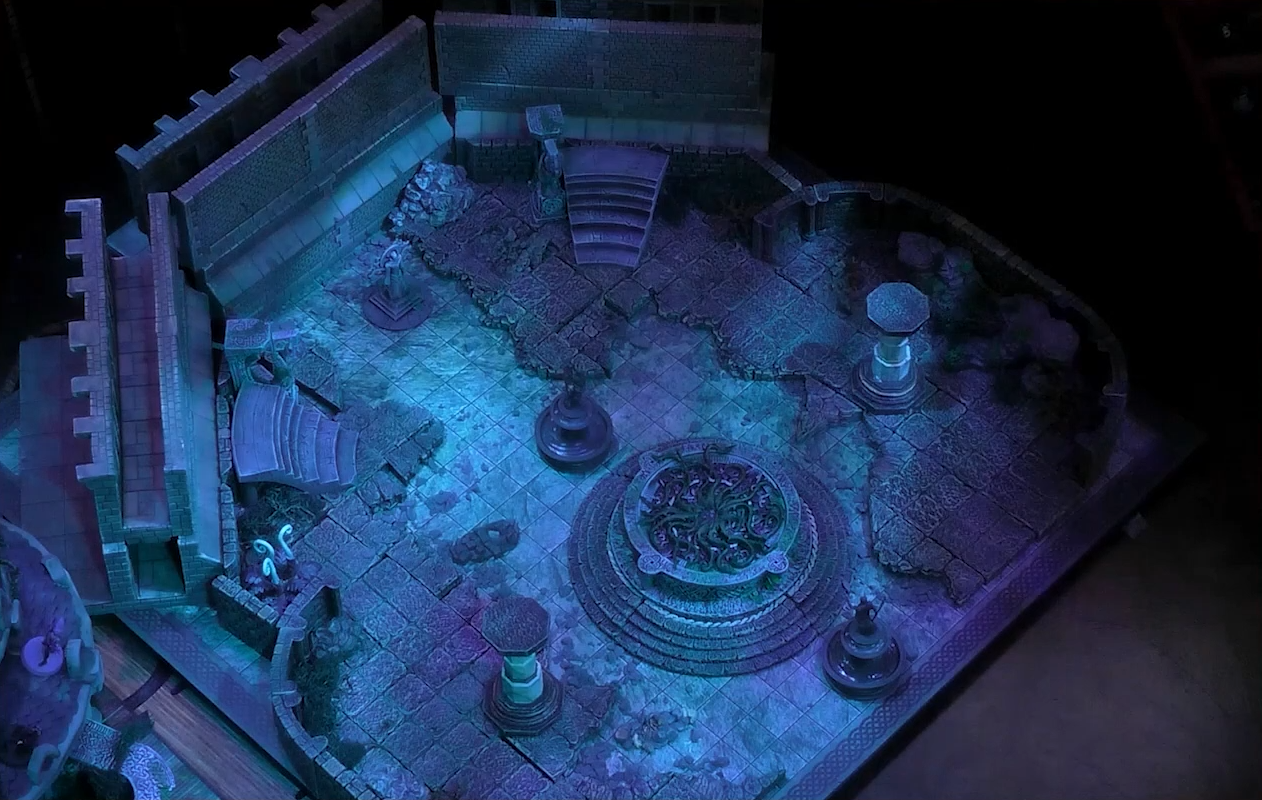 A battlemap of a chamber with a ruined stone floor, a vine-filled basin in the center, and two pillars on either end of it. Two statues stand in the front and rear of the chamber and stairs lead up on the sides. A lone white fiddlehead-like plant sits off in the corner.