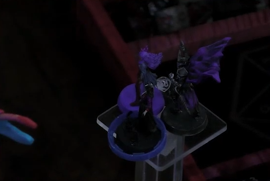 Miniatures for Imogen, Laudna, and Sorrowlord Zathuda on a raised plastic transparent platform. Sorrowlord Zathuda is wearing black armor and holding a sword aloft.