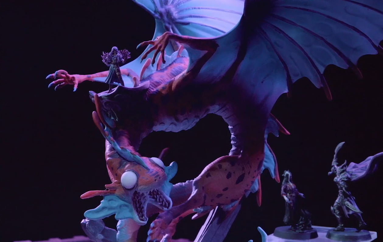 A miniature of Gloamglut, a massive snake-like dragon with wings outstretched. Imogen stands on its back. Her hands are palm-up in front of her, holding two roiling masses of purple energy. Laudna and Sorrowlord Zathuda are engaged in the foreground. The large, blank white eyes of Gloamglut’s fish-like head look forward, its split lower jaw open and filled with sharp teeth.