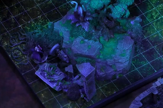 A battlemap focused on an outdoor area near some rocks and rubble. Dorian stands next to the unconscious Unseelie emissary. A cluster of white fiddlehead-like plants stands behond some bushes on nearby rocks.