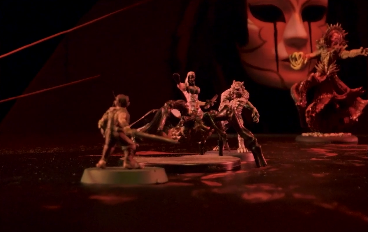 A battlemap with a glowing red blood floor focused on miniatures for Orym, Opal in spider form, and Chetney in wolf form. Opal stands between Orym and Chetney, a humanoid torso emerging from the abdomen of a giant spider. She holds her right hand out and carries a dagger in her left hand. In the background is Vespin Chloras, standing in front of the giant porcelain mask of the Matron.