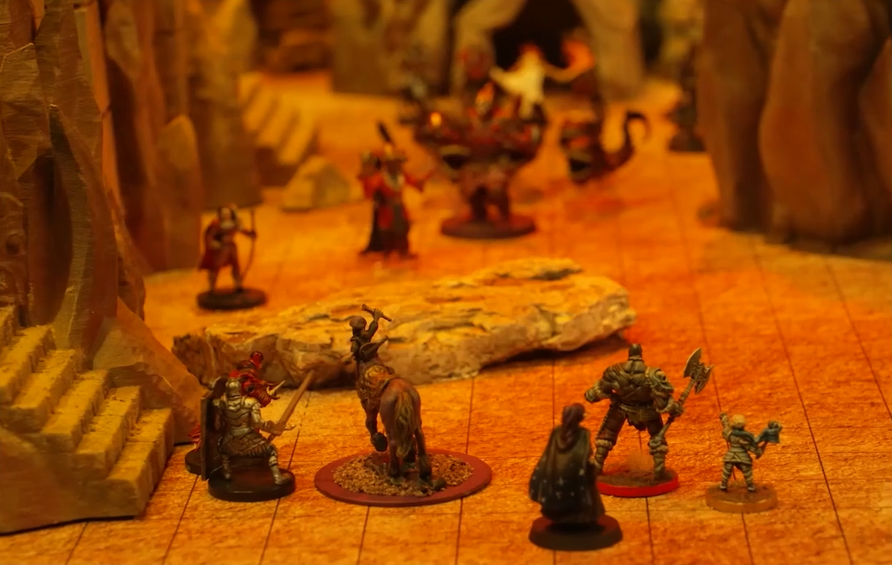 A battlemap focusing on Scanlan as a centaur, Vex, Grog, and Pike facing off against several enemies. They stand on a sand-covered ground at the base of a staircase leading up a stone wall.