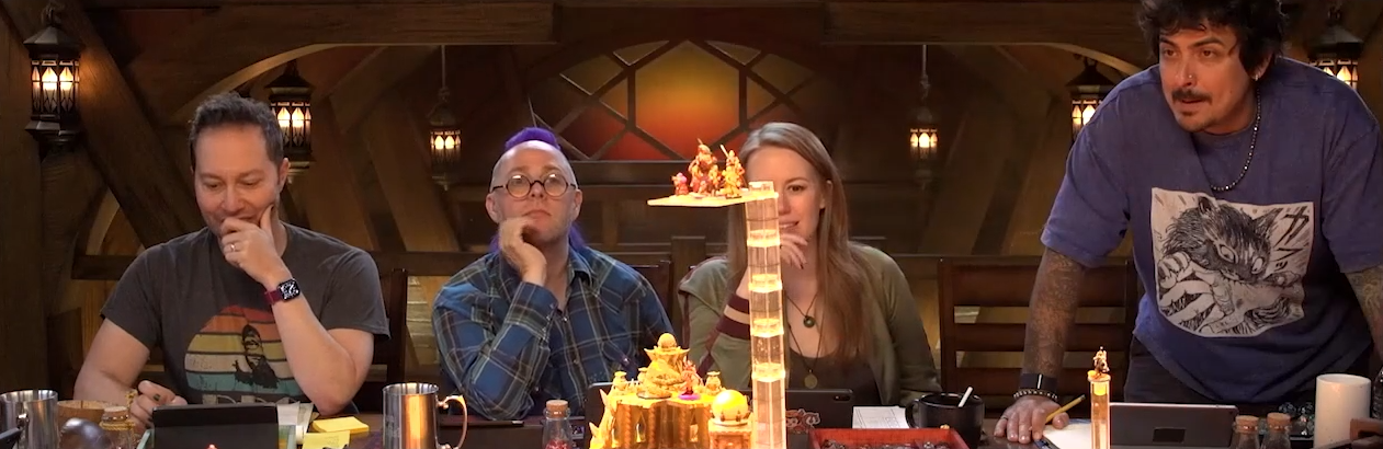 Sam, Taliesin, Marisha, and Robbie sit at the table. A transparent plastic platform with several miniatures placed on top of it is visible in the foreground.