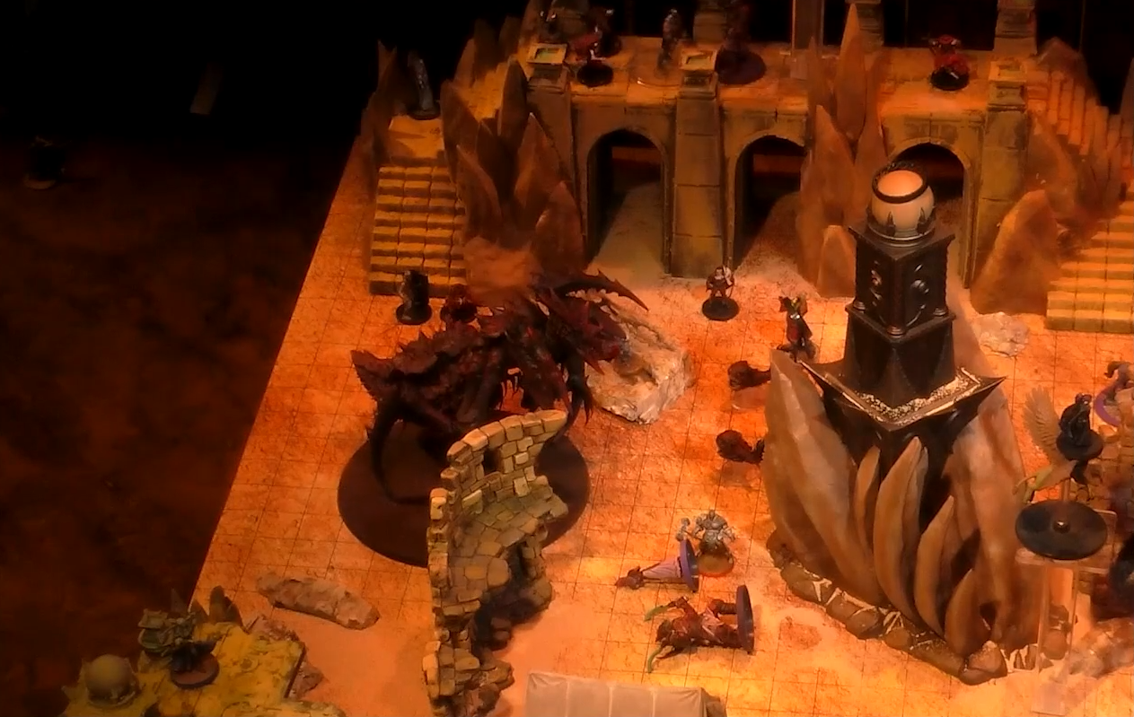 A battlemap of a large outdoor space filled with stone walls and parts of buildings. In the center is a tower with a pale white globe at the top. A number of combatants fill the area on the ground and atop the walls. A large, red, insectoid vidulch stands off to the side.