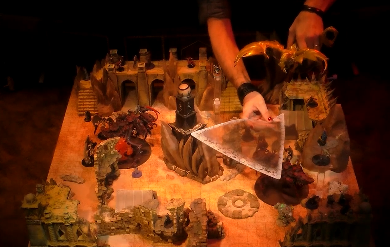 A battlemap of a large outdoor space filled with stone walls and parts of buildings. In the center is a tower with a pale white globe at the top. A number of combatants fill the area on the ground and atop the walls. Two vidulches stand on either side of the battlefield. Matt holds the miniature for a large brass dragon and measures the area of its breath weapon using a triangle-shaped sheet of plastic.