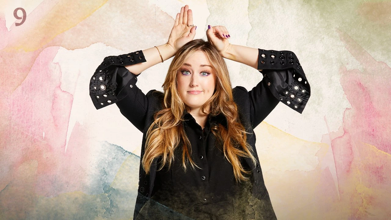 Thumbnail for episode 9. Ashley looks towards the camera with a subtle smile, her hands posed like long ears on top of her head. Her left hand is partially closed. She wears a black button-up shirt and stands against a colorful backdrop with splashes of greens, yellows, reds, and blues over white.