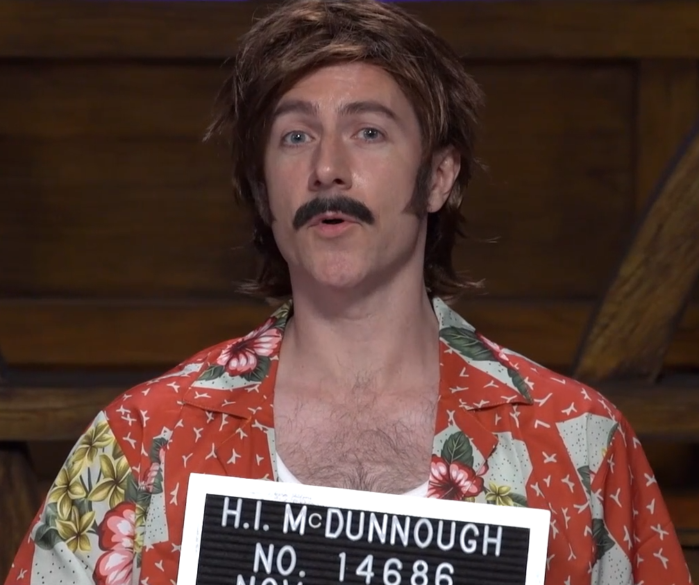 Matt with a brown wig and black mustache, wearing a bright orange floral button-up shirt. He holds in front of him a mug shot booking sign reading “H.I. McDUNNOUGH NO. 14686”.