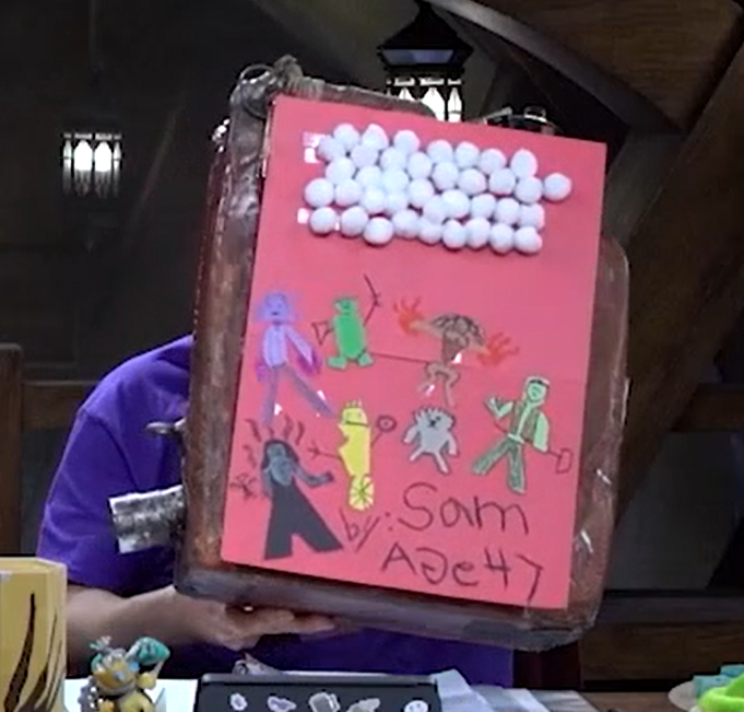 A rusty gas can with a page of red construction paper on it. A number of small cotton balls are stuck to the top of the paper to form a crude cloud. The members of Bell’s Hells are arranged below, made of other colors of construction paper in an artistic style reminiscent of a small child. In marker at the bottom is the text “by: Sam AGe 47”. The “G” in “age” is backwards.