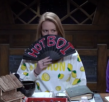 Marisha holding a black hand fan reading “HOT BOI” in red text outlined in white. The fan is covering the lower half of her face and she is peering over it. She is wearing a white sweatshirt with lemons on it and yellow text reading “LEMON”.