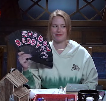 Marisha holding a black hand fan reading “SHADOW DADDY???” in red text outlined in white. She is wearing a pale green and white hoodie and smirking while glancing to her left.