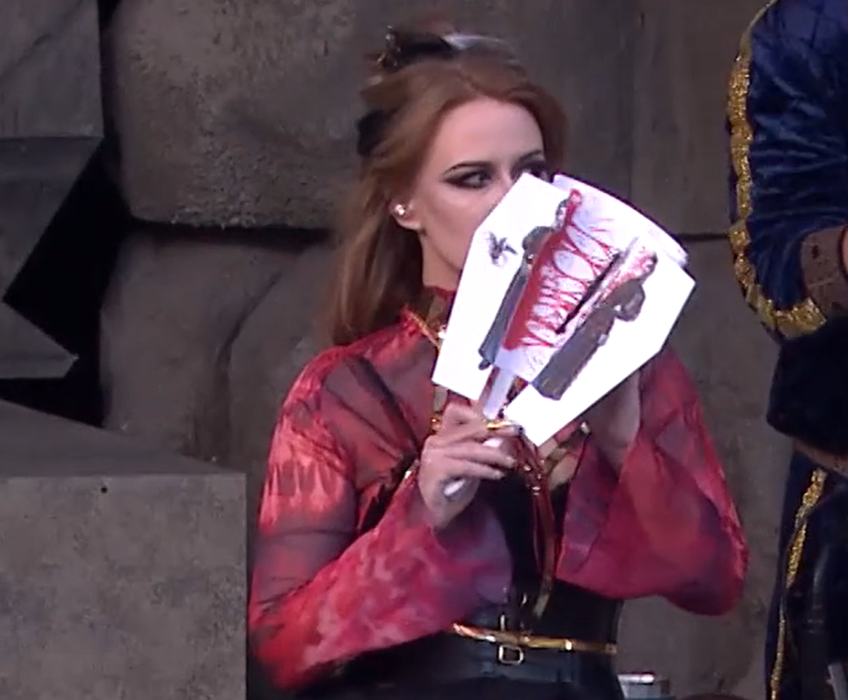 Marisha holding a partially open white paper hand fan with an image of Laudna split in half vertically and Pâté on the front. Strands of red blood connect Laudna’s two halves. Marisha is wearing a black dress with red sleeves and gold highlights, glancing to the side.