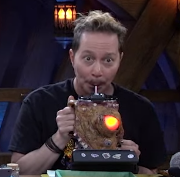 A tall cup covered in leathery monstrous skin. Its bulging glass eye is glowing orange. Sam is sipping from a straw placed in its top. His eyes are wide.