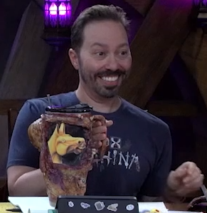 A tall cup covered in leathery monstrous skin. On the side is an image of the yellow carousel horse from “We Live in Time” with a goofy expression on its face. Sam is holding the cup’s handle and giving a toothy grin.