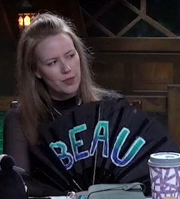 Marisha holding a black hand fan reading “BEAU” in green and blue lettering outlined in white. She is looking to the side seriously, her lips slightly pursed.