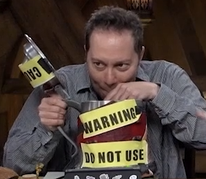 A comically-large silver beer stein with yellow labels “WARNING” and “DO NOT USE” on the side over red tape. A yellow label beginning “CAU” is taped on the opened lid. Sam drinks out of the stein using a straw.