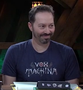 Sam wearing a blue T-shirt that says “VOX MACHINA”. The letters are stylized, where for example the “O” is a d20, the “X” is an hourglass, the “M” is a bow and arrow, and the final “A” is an axe.