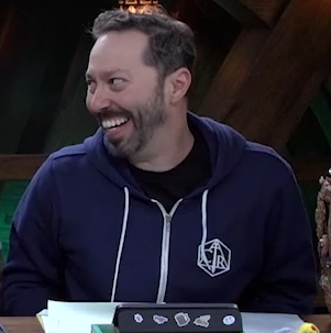 Sam wearing a dark navy hoodie. On the left chest in white is the Critical Role logo, a hexagon inscribed with a triangle (a 2D representation of a d20), inside of which is a downward-facing sword and the letters “CR”. The hoodie has a white zipper and drawstrings. Sam is looking to his right and smiling.