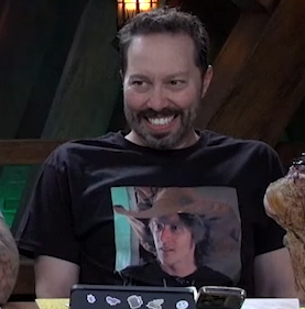 Sam wearing a black T-shirt with a photo of Matt printed on it. Matt is wearing a large brown wide-brimmed hat and looking into the middle distance with his mouth slightly open. Sam’s shoulders are raised and he is grimacing.