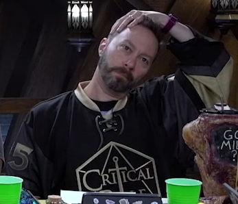 Sam wearing a Critical Role jersey. The jersey is mostly black with golden sleeves and collar. The large number “5” is visible on the right sleeve. The critical role logo is on the front. The logo is a hexagon inscribed with triangles (a 2D representation of a d20) inside of which is a downward-facing sword and the text “CRITICAL ROLE”, where the sword forms the T. Sam has his hand on his head, stretching his neck to the side.