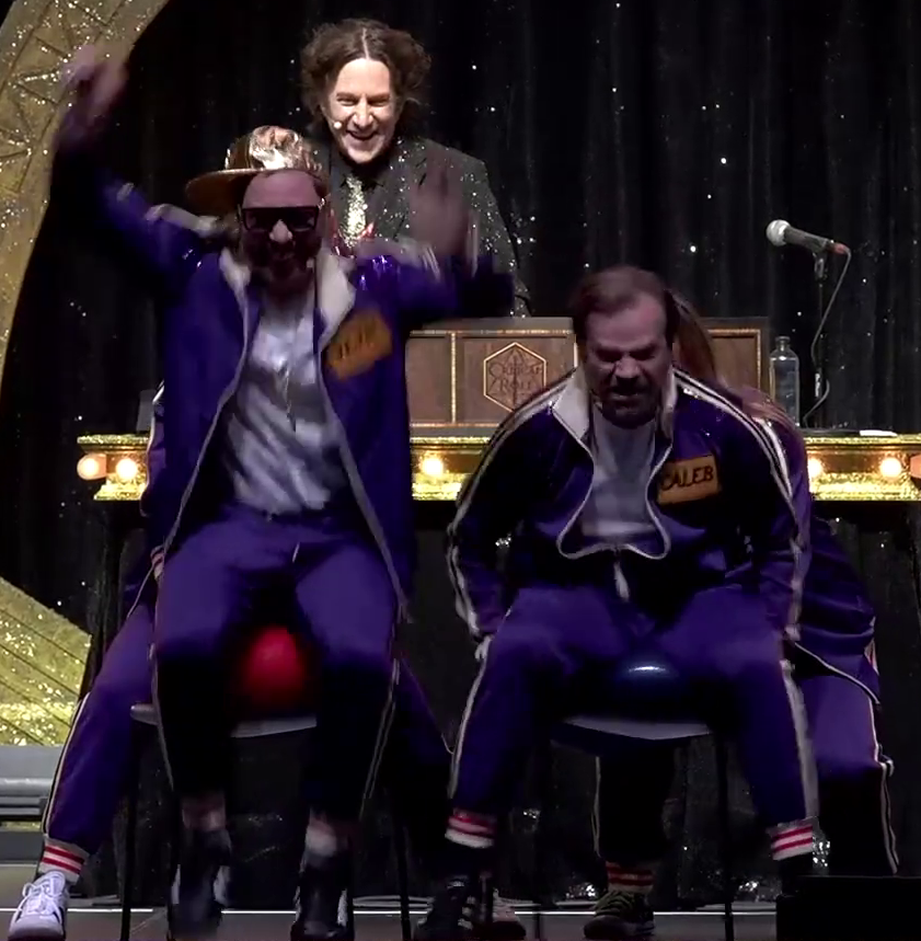 Sam and Liam on a stage in purple track suits, aggressively trying to burst large balloons by sitting on them on plastic chairs. Sam is smiling, bouncing high above his balloon. Liam is grimacing as he squishes his. Laura is behind Sam’s chair and Ashley is behind Liam’s, holding them. Matt stands in the background, laughing gleefully.