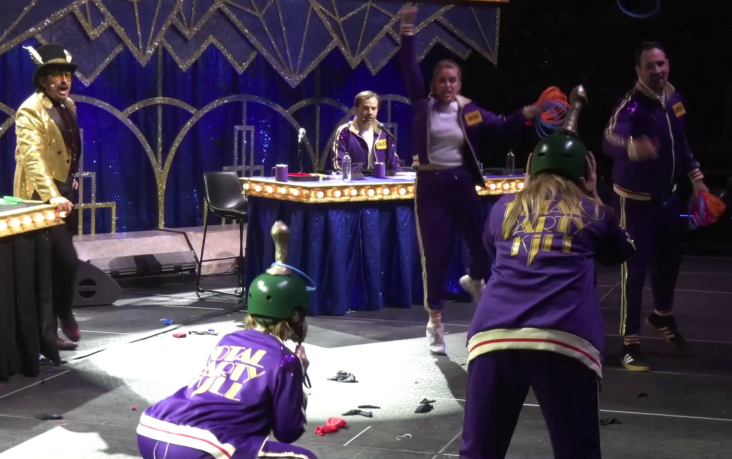 Marisha and Travis on a stage, tossing rings onto golden penis-shaped rods attached to helmets worn by Laura and Ashley. Liam observes from a table, while Robbie Daymond in a golden suit stares, his mouth agape. Marisha celebrates as she just scored a ring, while Travis watches as his next ring flies towards Ashley.